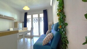 Ein modernes Wohnzimmer mit einem blauen Sofa, einem Essbereich mit weißem Tisch und Stühlen sowie einer Küchenzeile mit weißer Arbeitsplatte und einer Pendelleuchte.