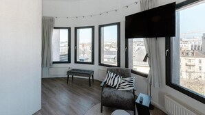 A modern living room with a curved ceiling, a large TV, a grey armchair with a striped pillow, a small table, and a bench.
