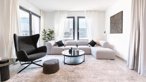 A modern living room with a grey armchair, a beige sofa set, a round coffee table, and a large window with curtains.