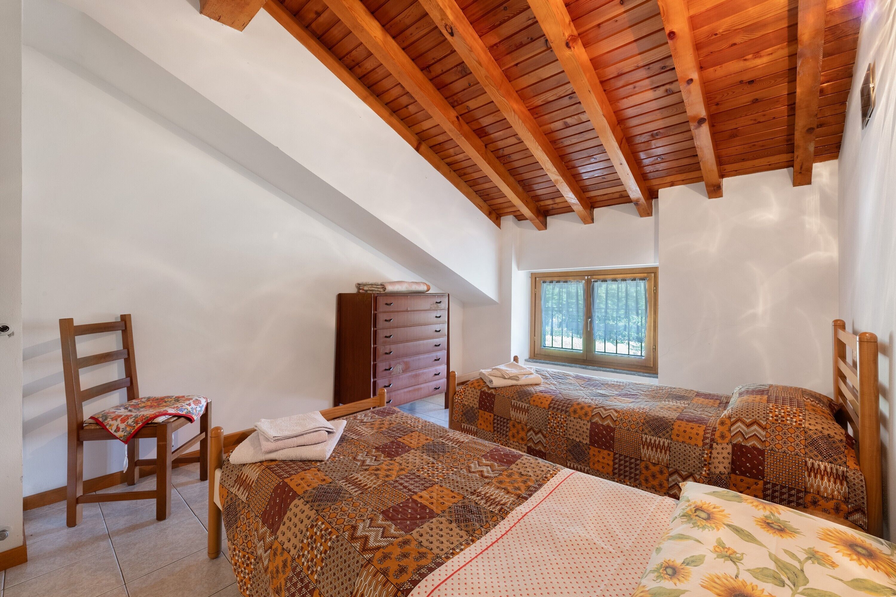 Quarto com duas camas, uma cadeira de madeira, cômoda e janela.