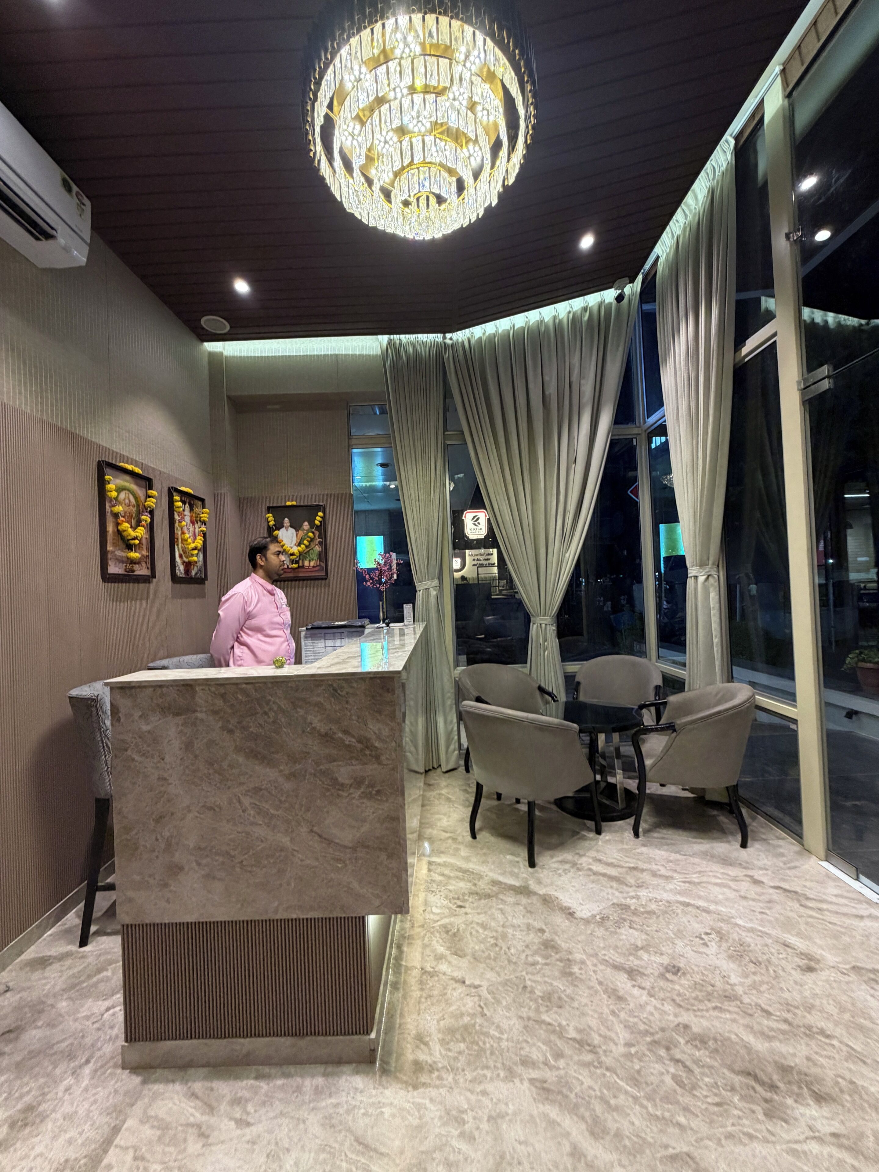 A hotel reception area with a marble counter, a chandelier, a seating area with chairs, and a large window with curtains.