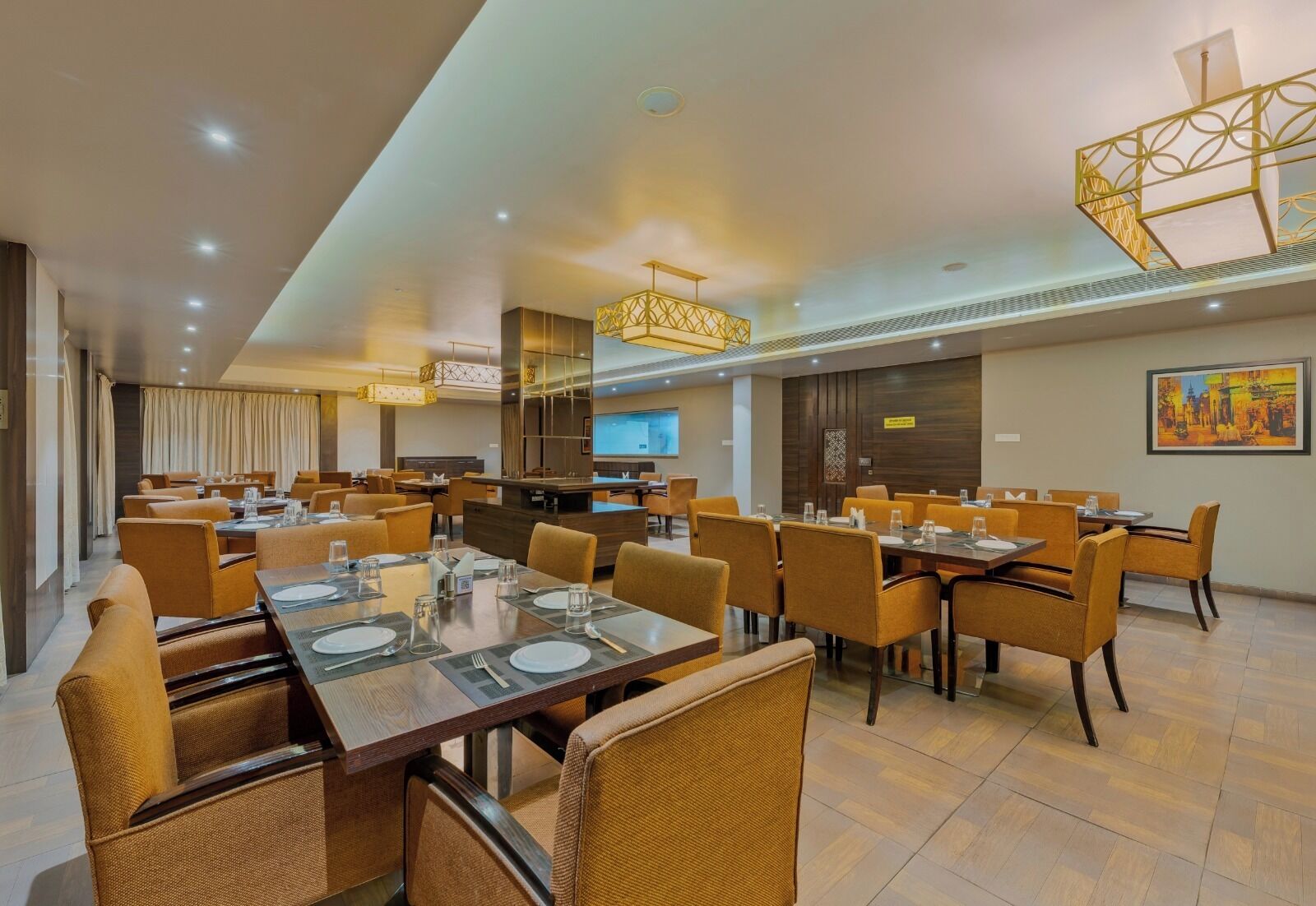 A modern hotel dining area with multiple tables and chairs, a large chandelier, and a television screen on the wall.