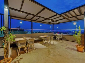 Una terraza en la azotea con asientos y mesas, ventanas amplias que ofrecen vistas a la ciudad y plantas en macetas.