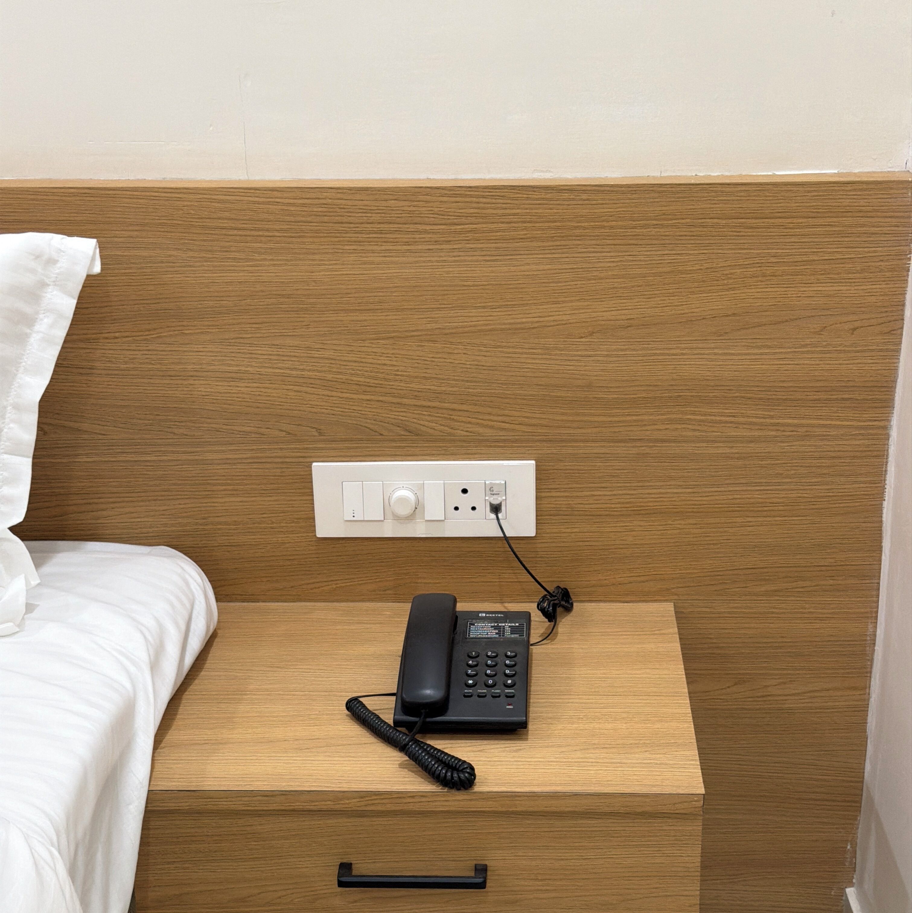 A wooden bedside table with a telephone, a power outlet, and a wall socket.