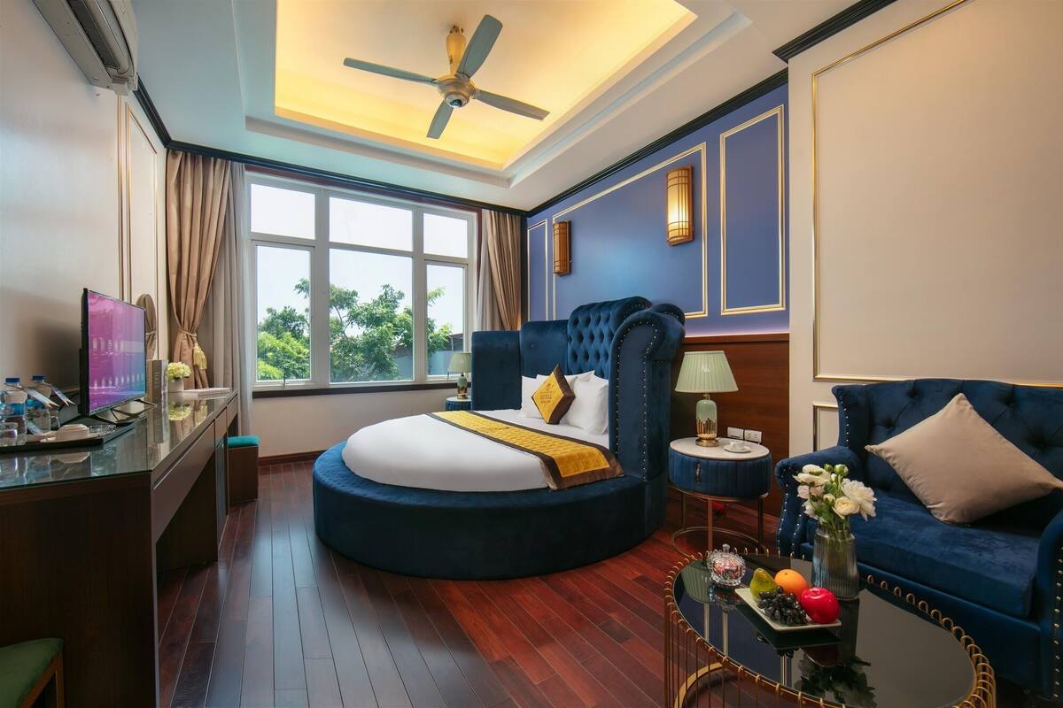 Habitación de hotel con una cama curvada, un escritorio con televisor, un sofá y una mesa de comedor con fruta.