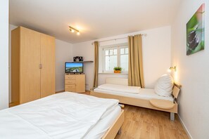 Un dormitorio con cama, televisor, armario, ventana con cortinas y mesita de noche con lámpara.