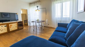 A living room with a blue sofa, a dining table with chairs, a TV on a cabinet, and a window with curtains. - SkyAttic - 801 by Houset (Milano)
