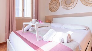 A neatly made bed with white linens, a pink blanket, and rolled towels. There is a small white table with a cup and a pink object. The room has a window with curtains, and two wall decorations.