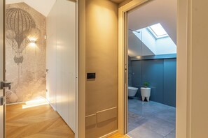 Um corredor moderno com piso de madeira, luminária fixada na parede e porta que dá para um banheiro com claraboia e uma pequena planta em vaso.
