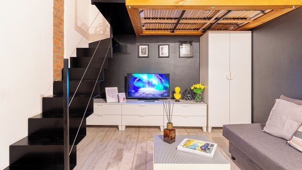 A modern living room with a black staircase, a flat-screen TV, a white sofa, and a wooden ceiling structure.