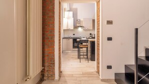 A narrow hallway leading to a kitchen with a brick column, a dining area with a table and chairs, and a black staircase.