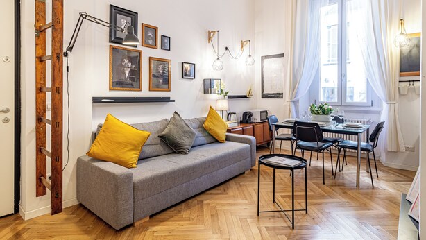 A modern living room with a grey sofa, yellow cushions, a wooden ladder, a dining table with chairs, and a large window with sheer curtains.