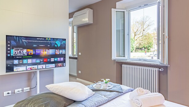 A hotel room with a flat-screen TV mounted on the wall displaying various streaming apps, a bed with a blue and white striped bedspread, a window with a view of trees and buildings, and a radiator below the window. - Isolino Bridge Apartment by Houset (Monza)