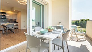 A modern dining area with a table and chairs, a kitchen in the background, and a view of greenery outside.
