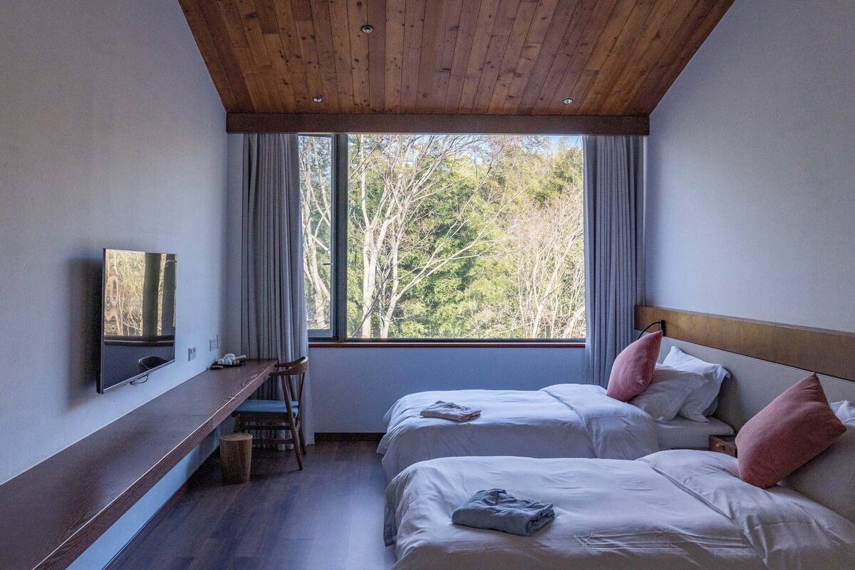 Un dormitorio con dos camas, un escritorio, un televisor y un ventanal con vistas a árboles.