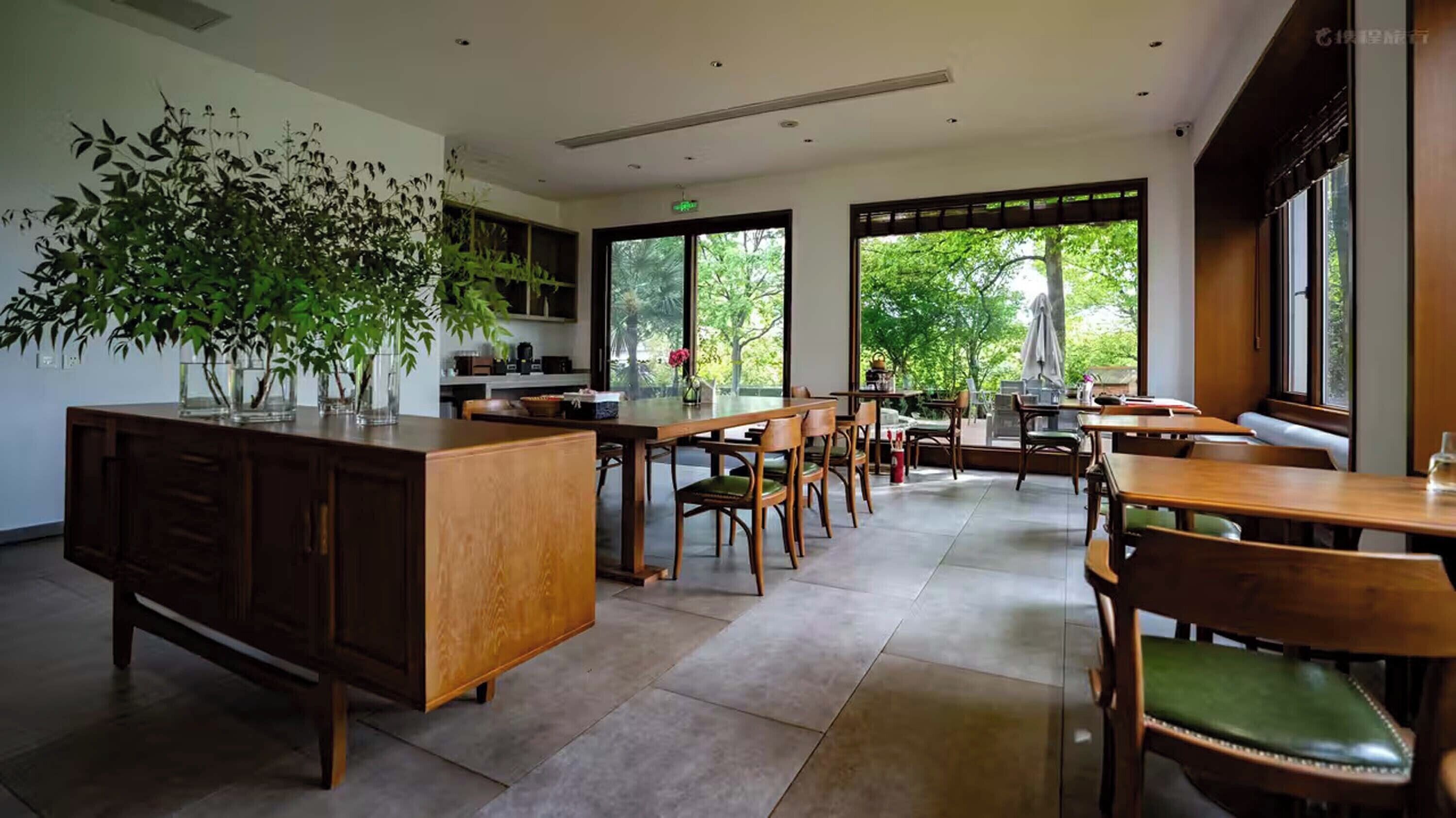 A spacious dining area with wooden tables and chairs, a large potted plant, and large windows offering a view of greenery outside.