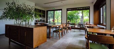 A spacious dining area with wooden tables and chairs, a large potted plant, and large windows offering a view of greenery outside.