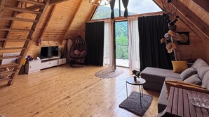A spacious loft bedroom with a sloped ceiling, wooden beams, a large window, a hanging chair, a sofa, and a TV. - Villa Cottage Merisi (Keda)