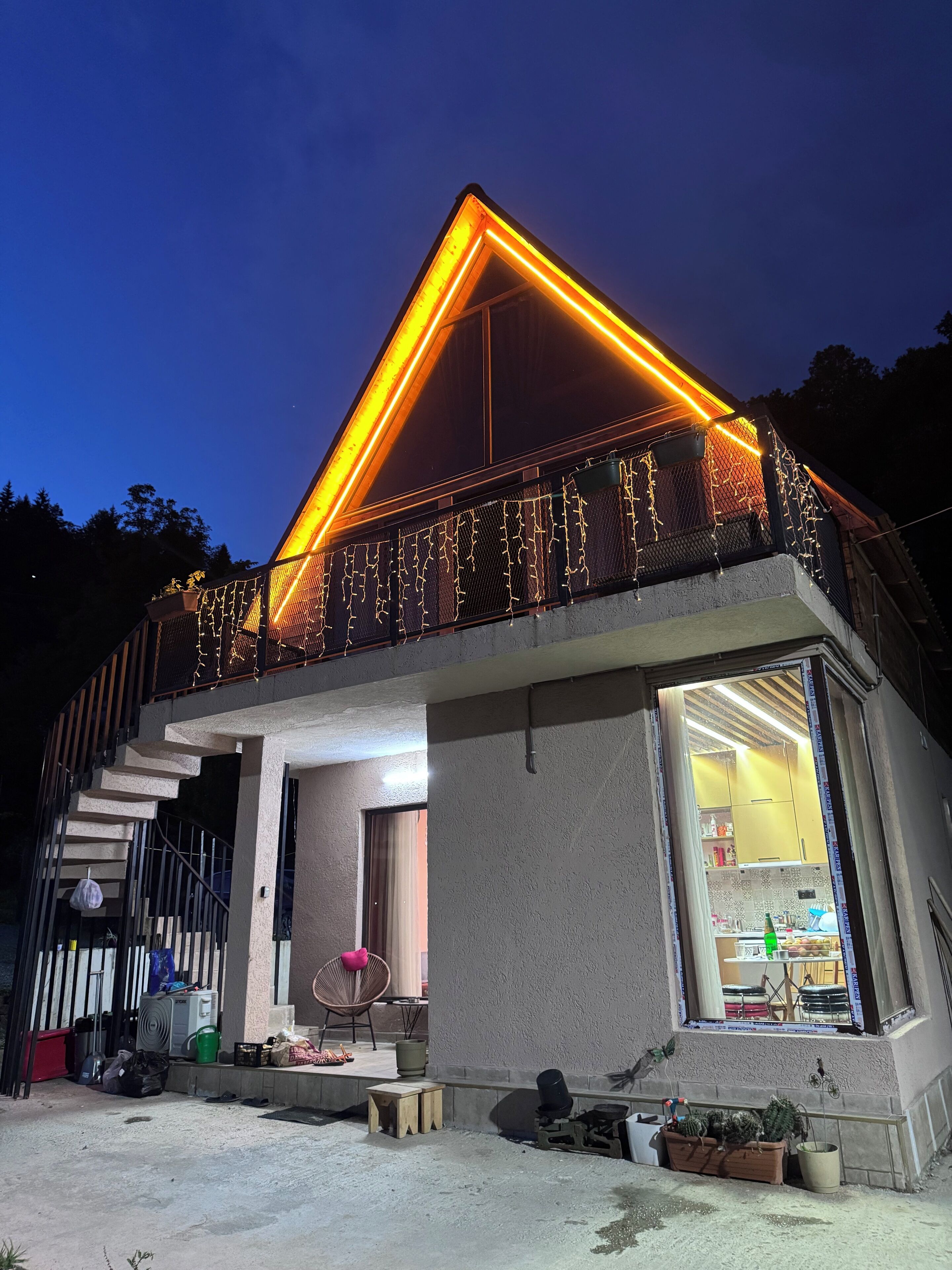 A modern house with a balcony decorated with string lights, a visible kitchen area, and a staircase leading to the upper floor.