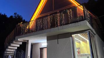 A modern house with a balcony decorated with string lights, a visible kitchen area, and a staircase leading to the upper floor.