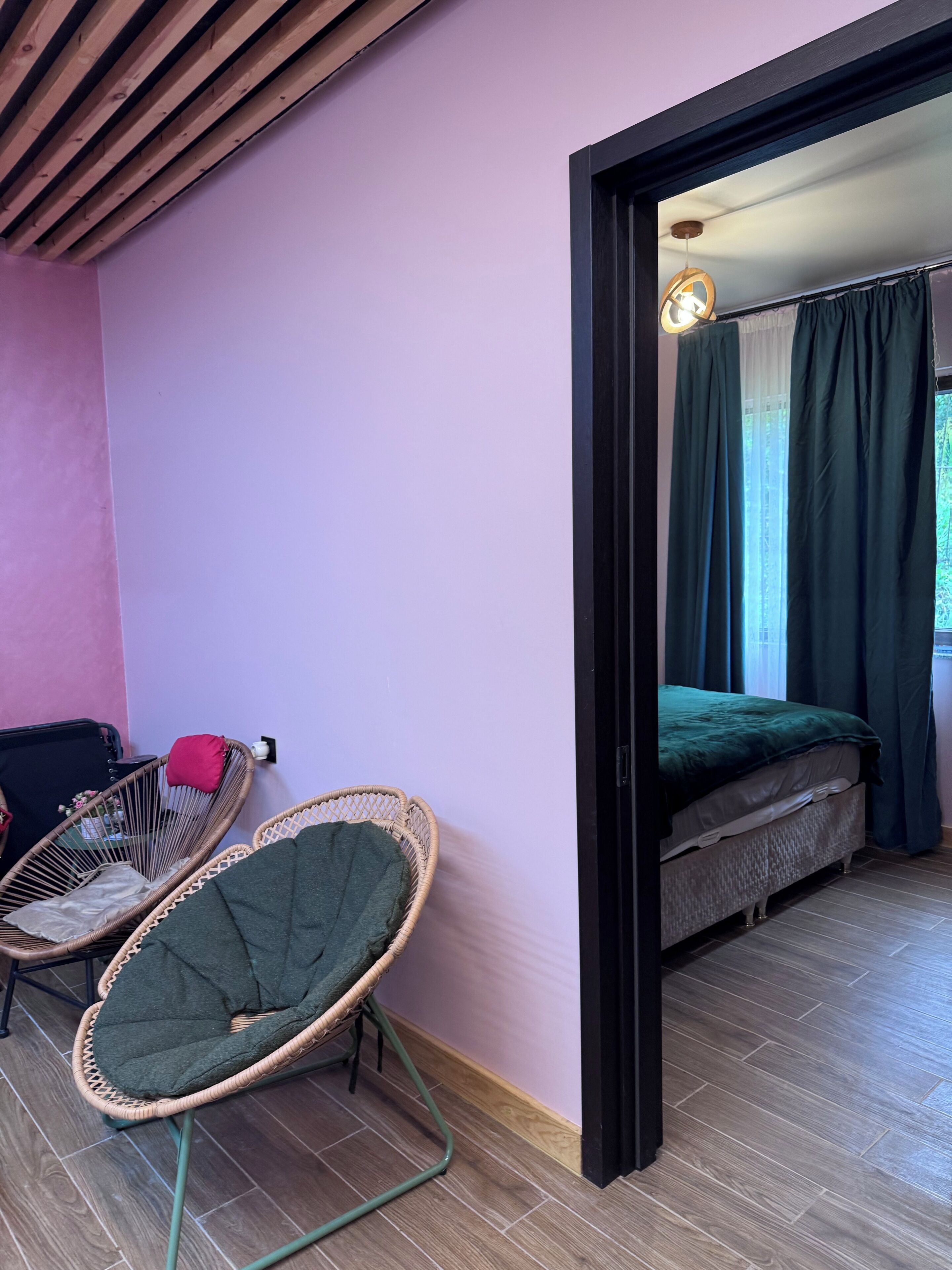 Ein Zimmer mit einem Bett, einem Korbstuhl und Blick ins Freie durch ein Fenster mit Vorhängen.