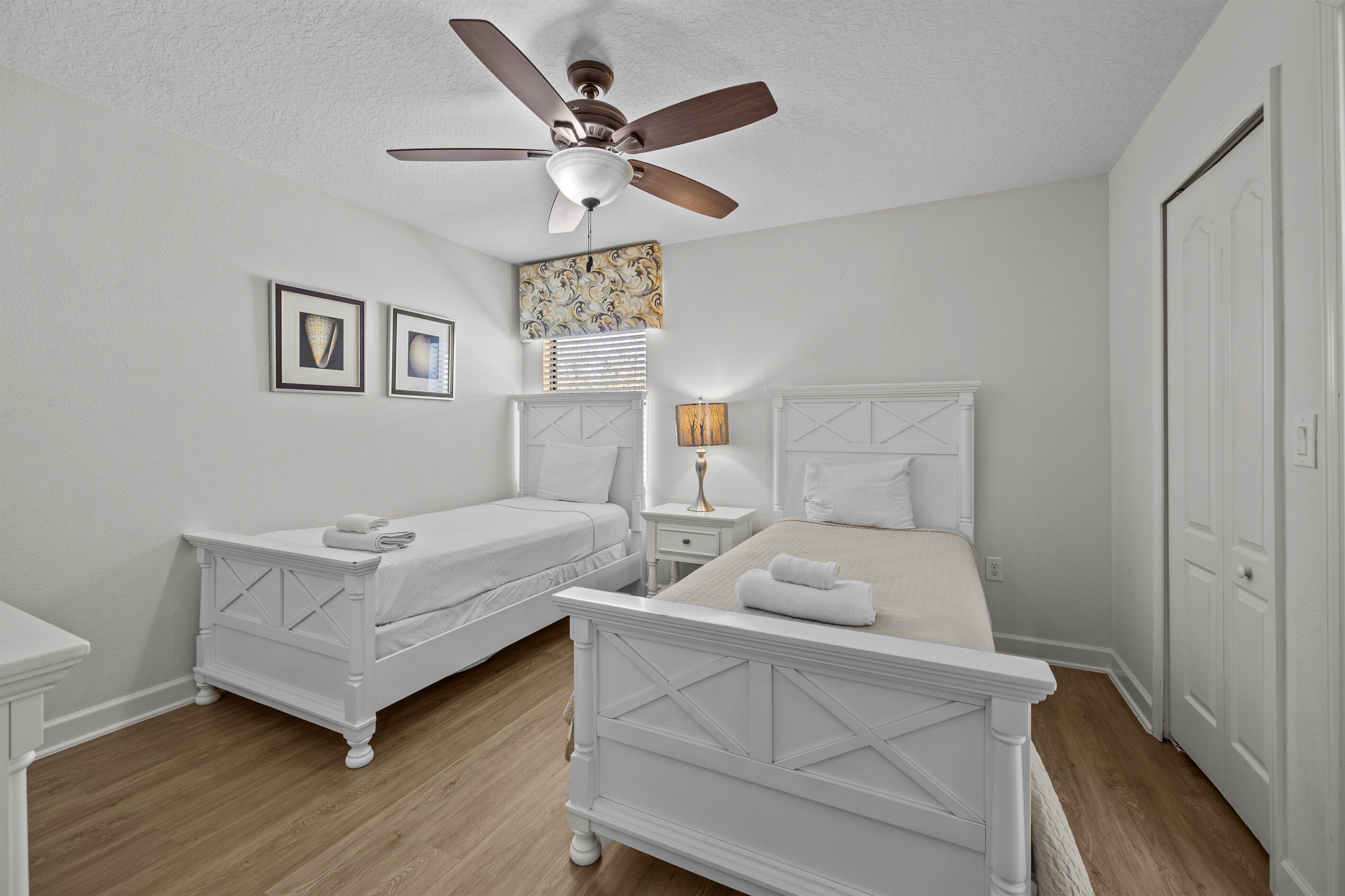 Une chambre à coucher avec deux lits blancs, un ventilateur de plafond, une table de chevet avec une lampe et une grande fenêtre avec des stores.