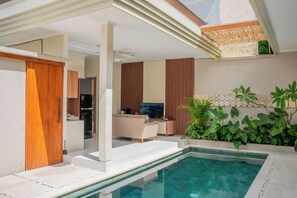 A modern indoor pool area with a clear view of the living room, a sofa, and a television. - Villa Kamandari (Seminyak)
