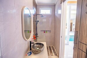 A bathroom with a stone sink, a mirror, a shower, and a small window. - Villa Kamandari (Seminyak)