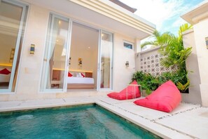 A modern hotel room with a large sliding glass door leading to a balcony, a small pool area with red cushions, and a tropical plant wall. - Villa Kamandari (Seminyak)