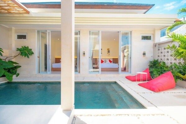 A modern villa with a swimming pool, glass doors leading to a bedroom, and a patio area with cushions. - Villa Kamandari (Seminyak)