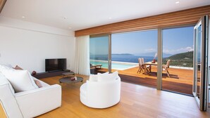 Ein modernes Wohnzimmer mit einem weißen Sofa, einem weißen Hocker und einem Fernseher. Große Glasfenstertüren führen zu einer Außenterrasse mit Pool und Meerblick.
