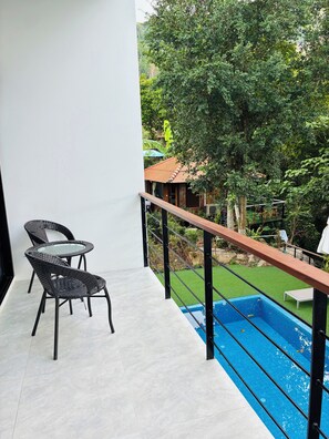 A balcony with two wicker chairs, a glass table, a pool, and a view of a garden and house. - Hillside Haven-Luxury Room (Koh Samui)