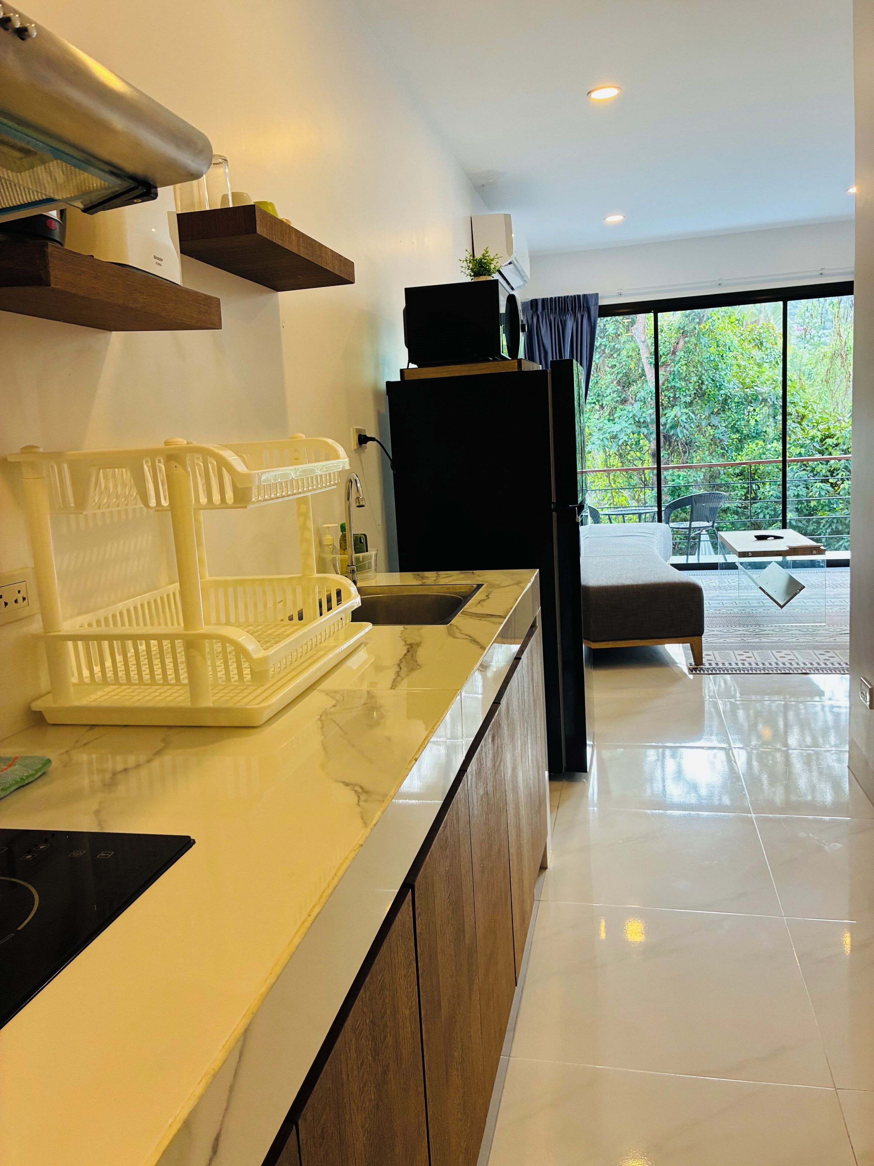 A modern kitchen with a marble countertop, a built-in oven, and a sink. There is a dining area with a table and chairs, and a view of greenery through large windows.