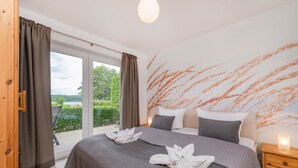 A bedroom with a large bed, a nightstand, a window with curtains, and a view of greenery outside.