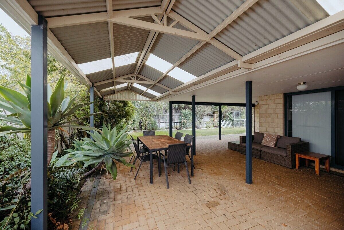 An outdoor covered patio area with a brick floor, a wooden table with chairs, a sofa, and a large plant.