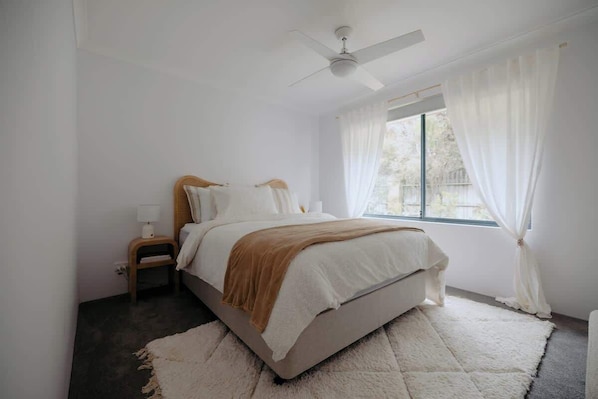 Ein Schlafzimmer mit einem Bett, einem Nachttisch, einem Fenster mit Vorhängen und einem Deckenventilator.