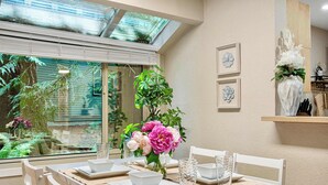 Ein Essbereich mit Tisch für vier Personen, ein großes Fenster mit Blick auf Grünflächen und eine Wandvase mit Blumen.