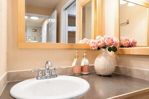 A bathroom with a white sink, a mirror, and a vase with pink flowers. There are two bottles of soap and a decorative vase on the counter. - @ Marbella Lane - Cozy Forest Cabin Min from DWNTN (Redmond)