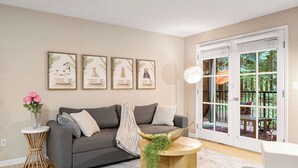 Ein Wohnzimmer mit grauem Sofa, einem runden Holztisch mit Pflanze, einem weißen Beistelltisch mit Blumenvase und einem großen Fenster mit Blick auf Grünflächen.
