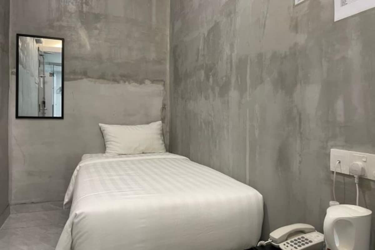 A small, single-bed room with a plain white bedspread, a white telephone, and a white wall-mounted toilet.