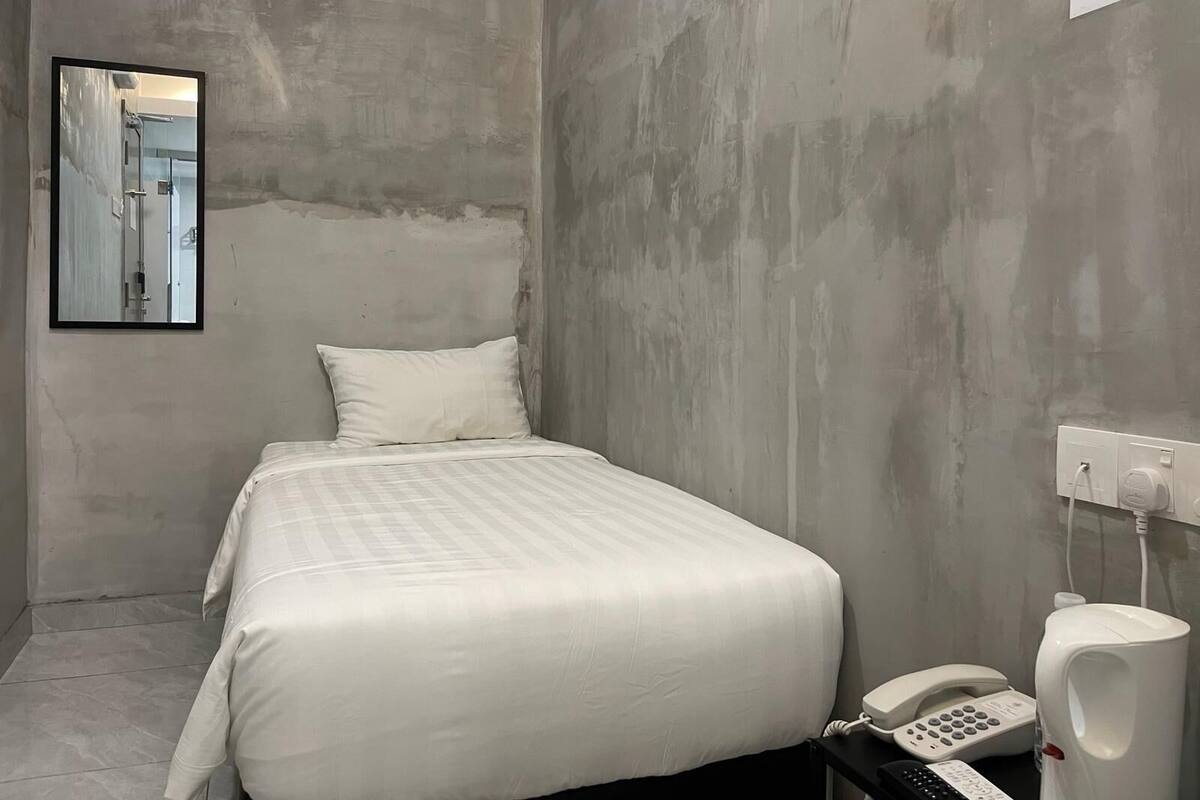 A small, single-bed room with a plain white bedspread, a white pillow, a bedside telephone, and a white urinal.