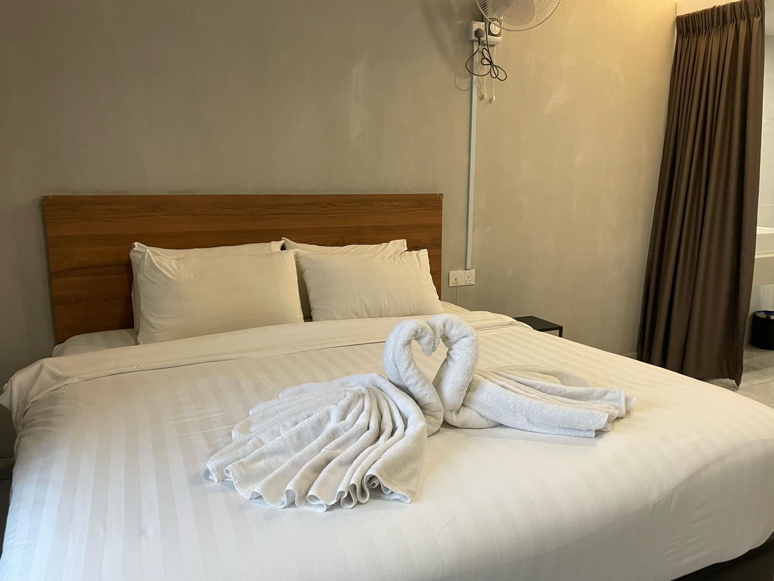 A hotel room with a large bed, white linens, a wooden headboard, a wall-mounted fan, and a brown curtain.