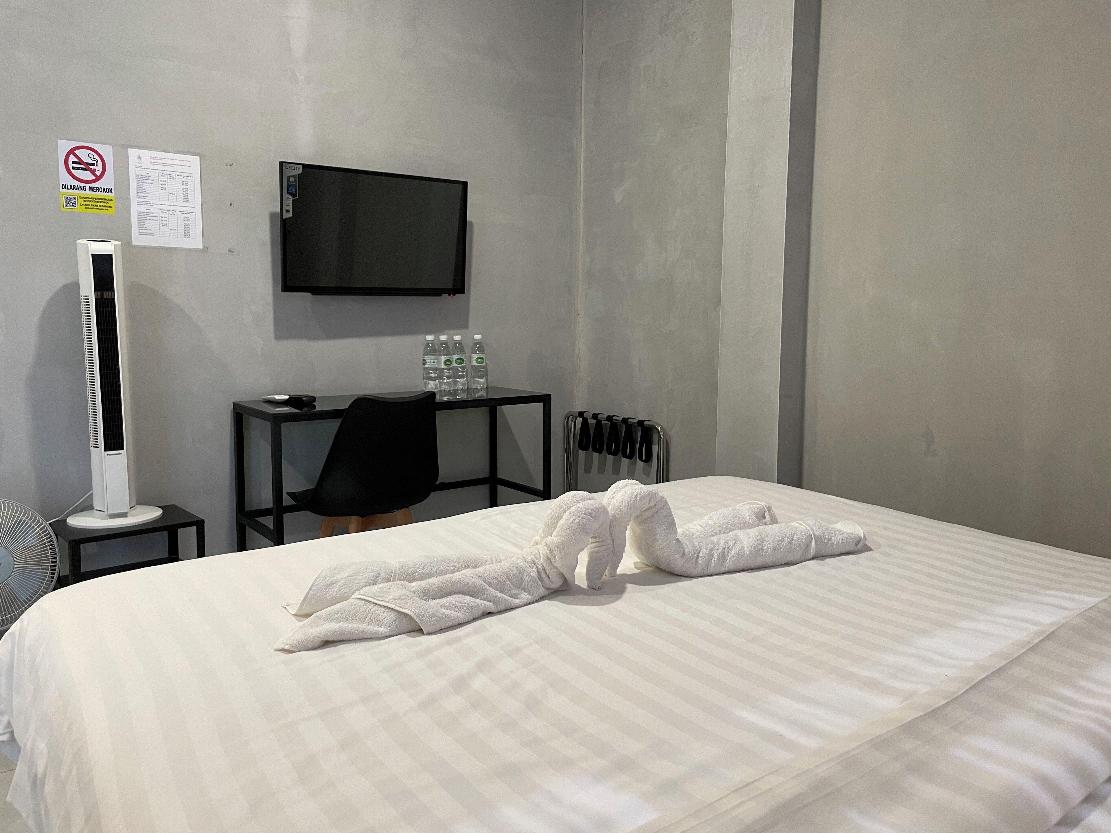 Ein Hotelzimmer mit Bett, Flachbildfernseher an der Wand, Schreibtisch mit Stuhl, Heizkörper und Ventilator.