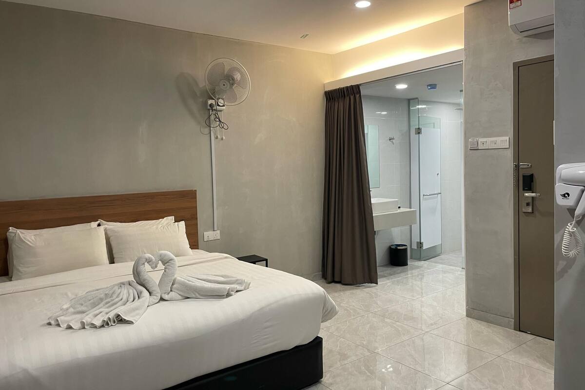 A hotel room with a large bed, a bedside table, a wall-mounted fan, a bathroom visible through an open door, and a telephone.