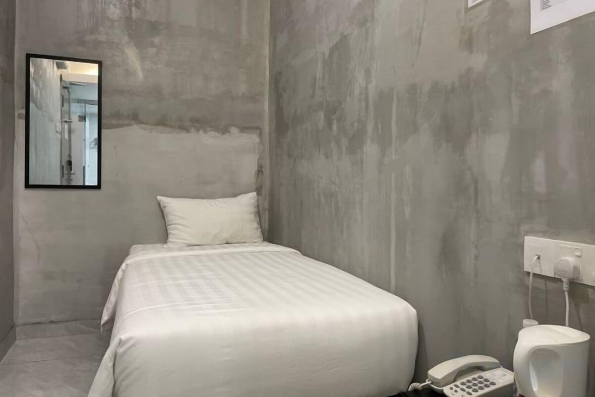 A small, single-bed room with a plain white bed, a white telephone, and a white urinal.