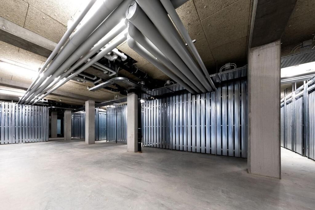 Un parking couvert avec des piliers en béton, des casiers métalliques et des tuyaux apparents au plafond.