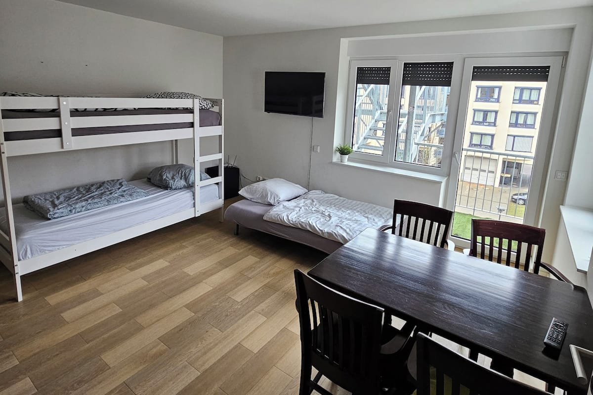 Et lille, moderne værelse med køjeseng, enkeltseng, spisebord med stole, et fjernsyn ophængt på væggen og et vindue med udsigt til bygninger.