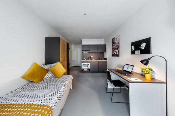 Una habitación compacta con cama, escritorio, silla, kitchenette pequeña y microondas.