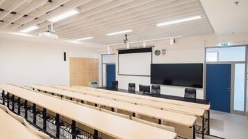 Un aula con filas de escritorios, un proyector, una pizarra y una puerta con señal de salida.
