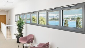 A modern interior with a row of windows offering a view of a body of water and greenery. There are two chairs and a small table in the foreground. - Akademis Fit Split (Split)